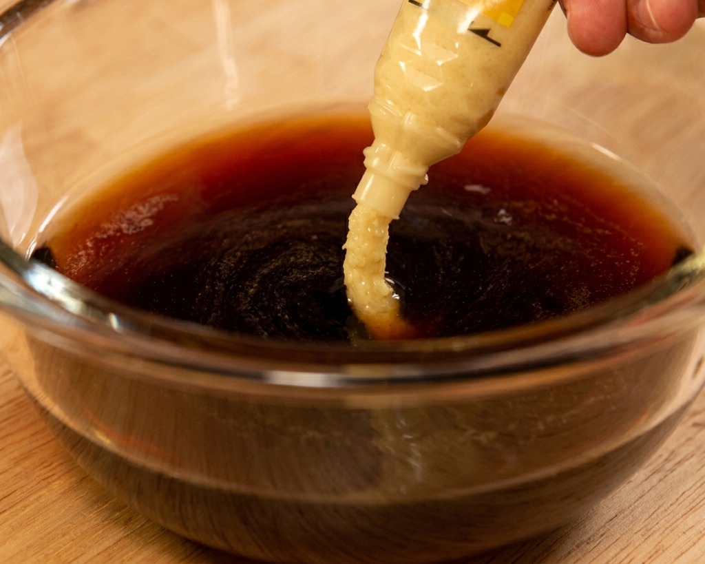 Grated ginger paste being squeezed from a tube into a dark soy-based sauce mixture in a glass bowl.