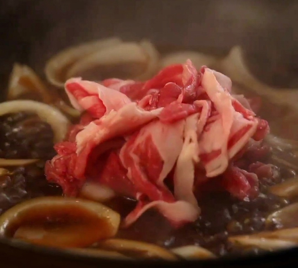 Raw beef belly slices being added to a simmering pan of onions and Japanese savory sauce.