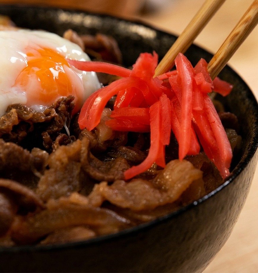 A close-up shot of wooden chopsticks placing bright red slivers of pickled ginger onto a steaming beef bowl topped with a soft-boiled egg.