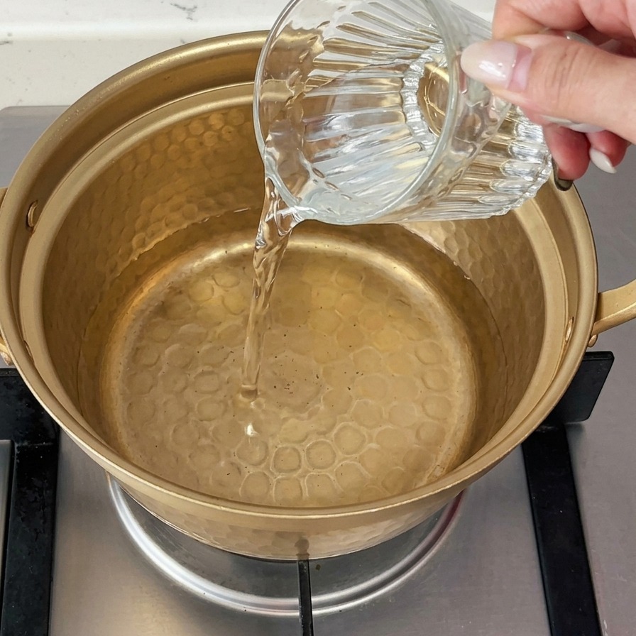 Cold water being poured from a glass cup into a small, gold-colored metal pot sitting on a gas stove.
