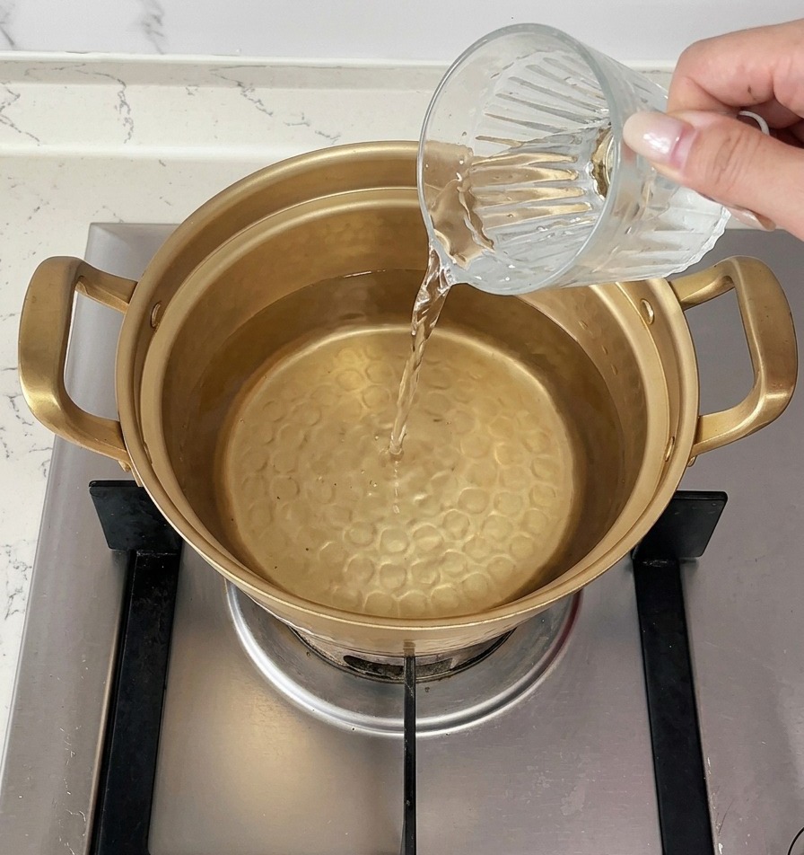 A hand pouring a cup of cold water into a gold pot that is already filled with hot water on the stove.
