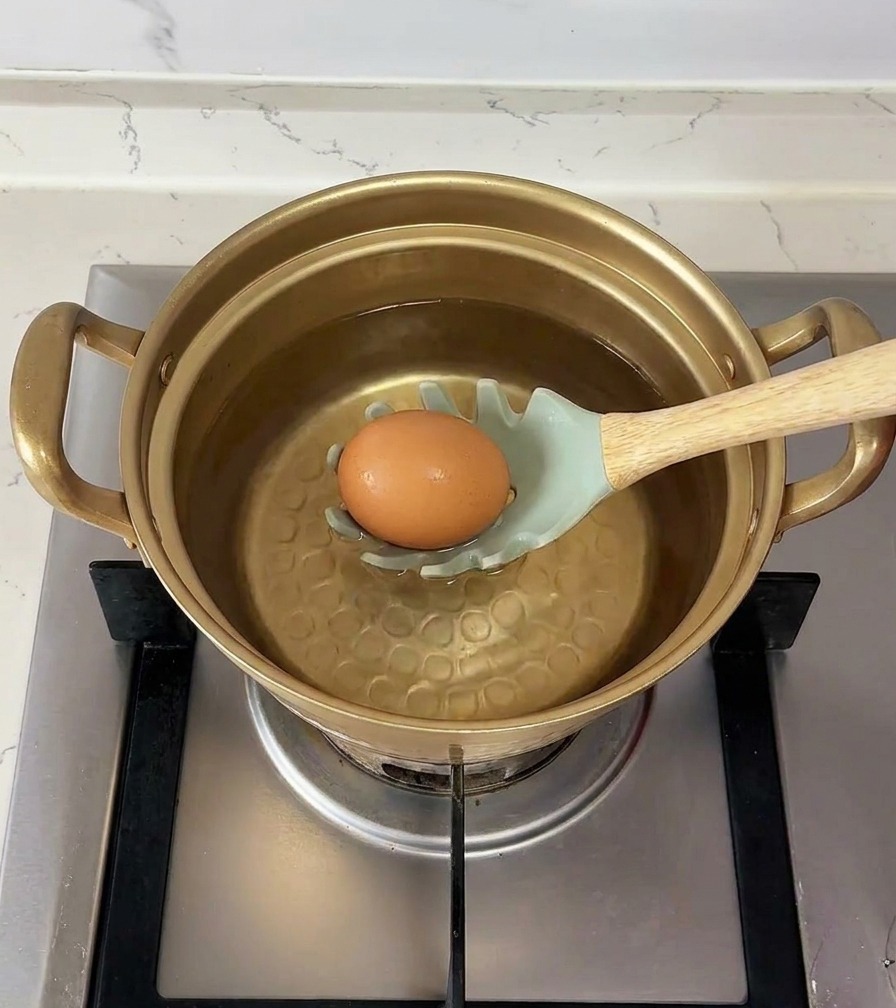 A brown egg resting on a light green slotted spoon being carefully lowered into a pot of water.