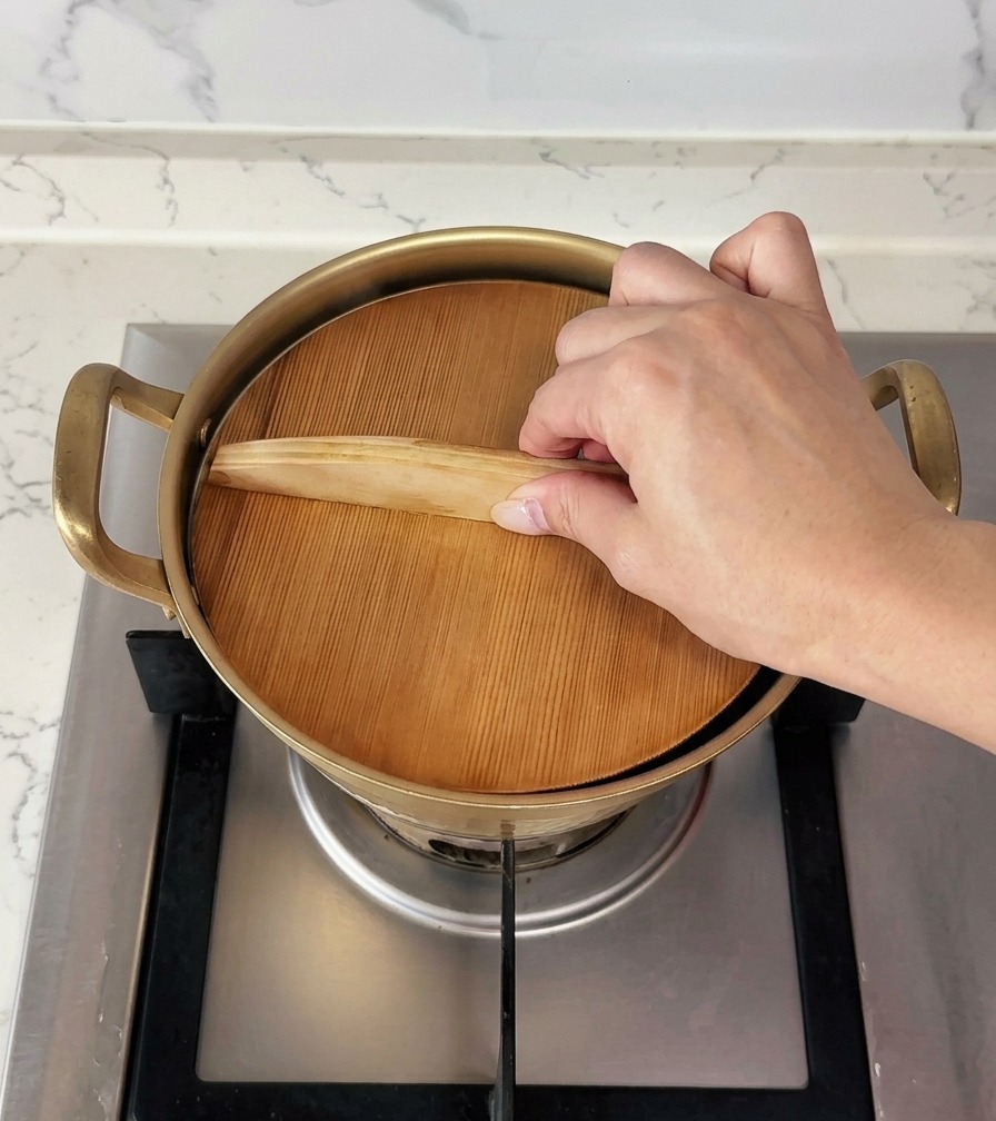 A hand placing a flat wooden lid onto a small gold cooking pot to cover it.