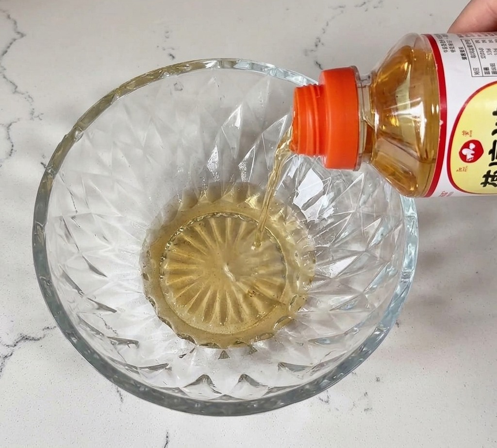 Clear mirin liquid being poured from a bottle into a crystal glass bowl on a white countertop.