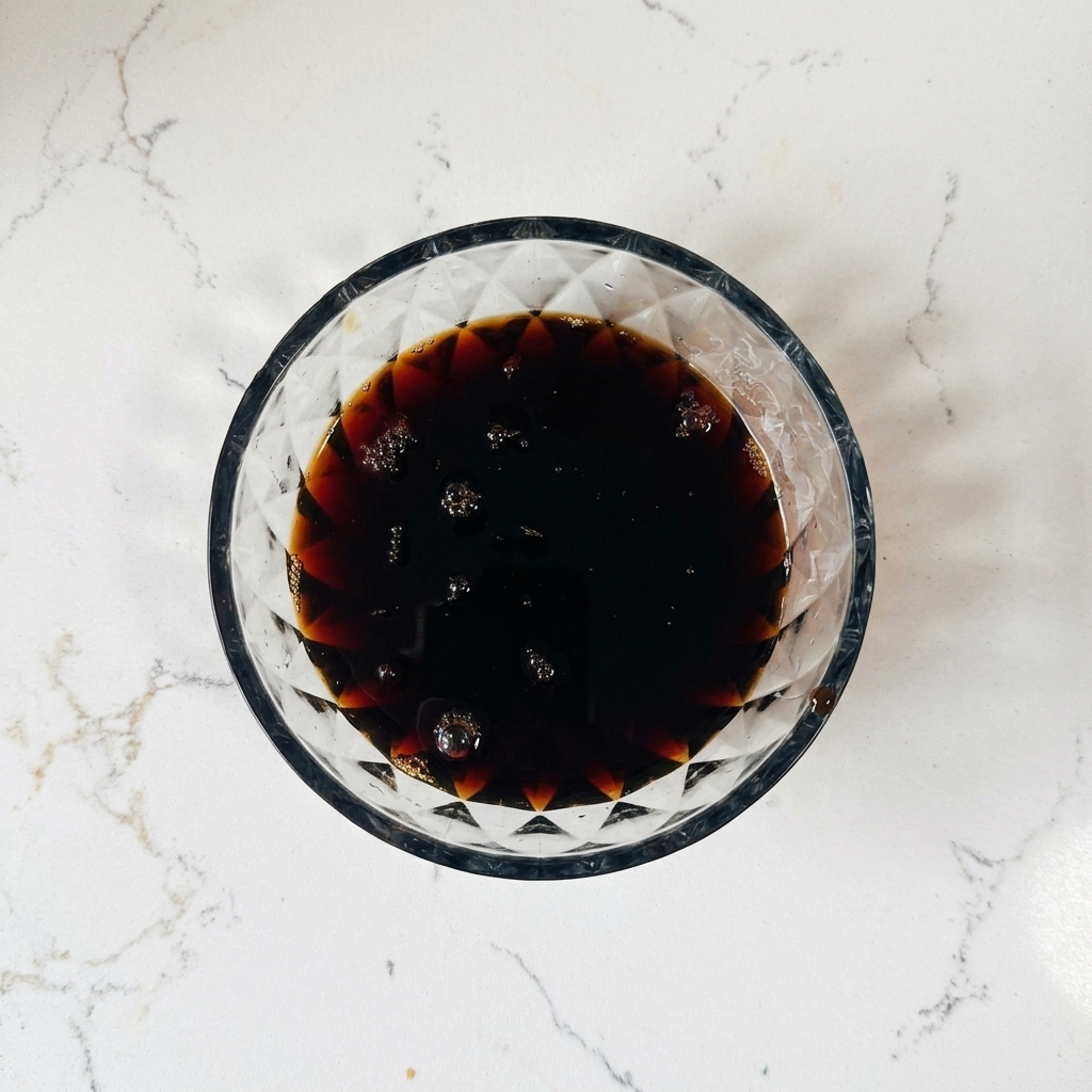 A top-down view of a glass bowl containing a dark brown liquid sauce with small air bubbles on the surface.