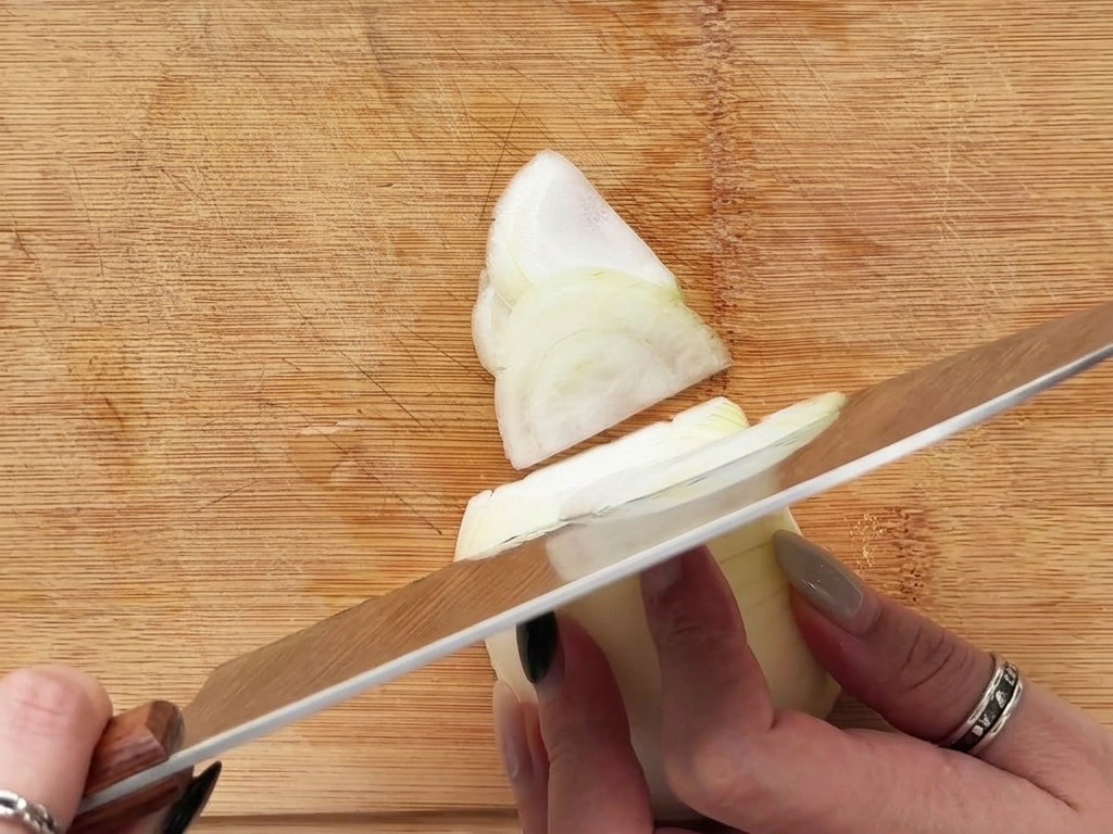 Hands slicing a white onion into thin crescent shapes on a wooden cutting board.