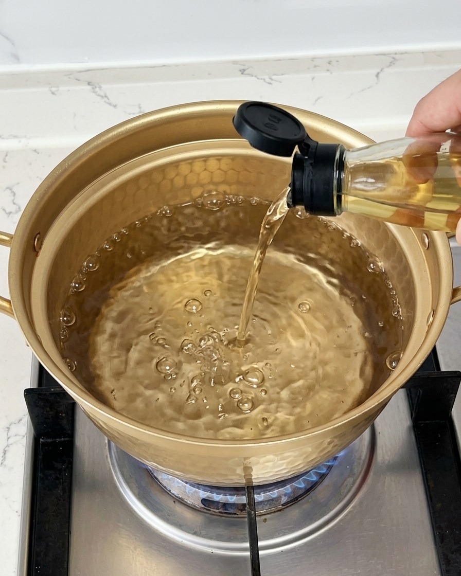 Pouring clear cooking wine from a glass bottle into a gold pot filled with boiling water.