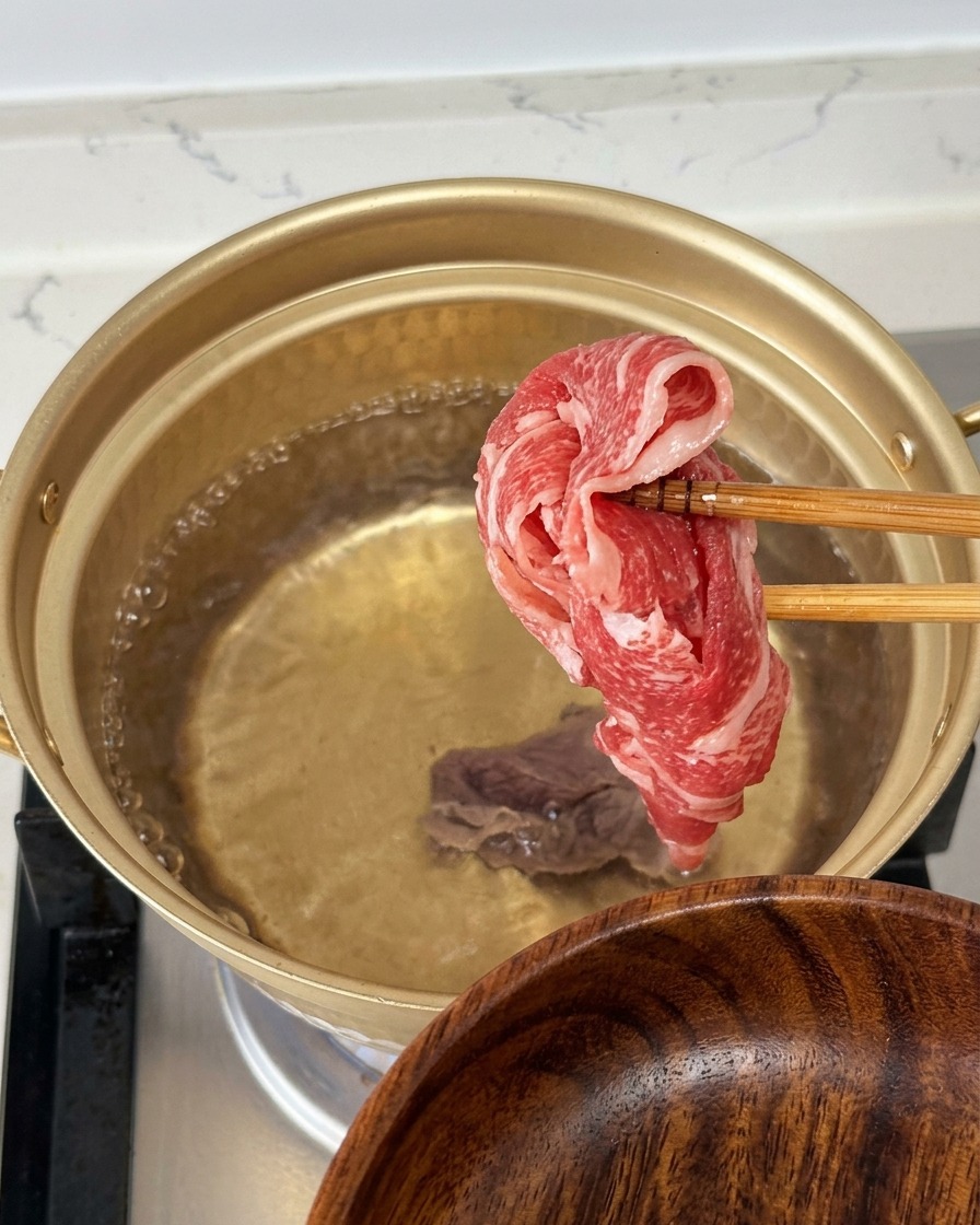 Holding a thin slice of raw, marbled beef with chopsticks over a gold pot of boiling water.