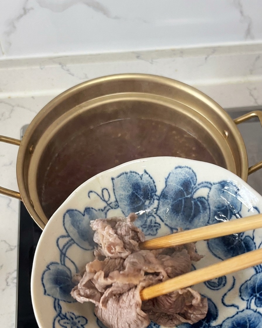 Lifting partially cooked, opaque beef slices out of a pot with chopsticks and transferring them to a blue and white patterned plate.