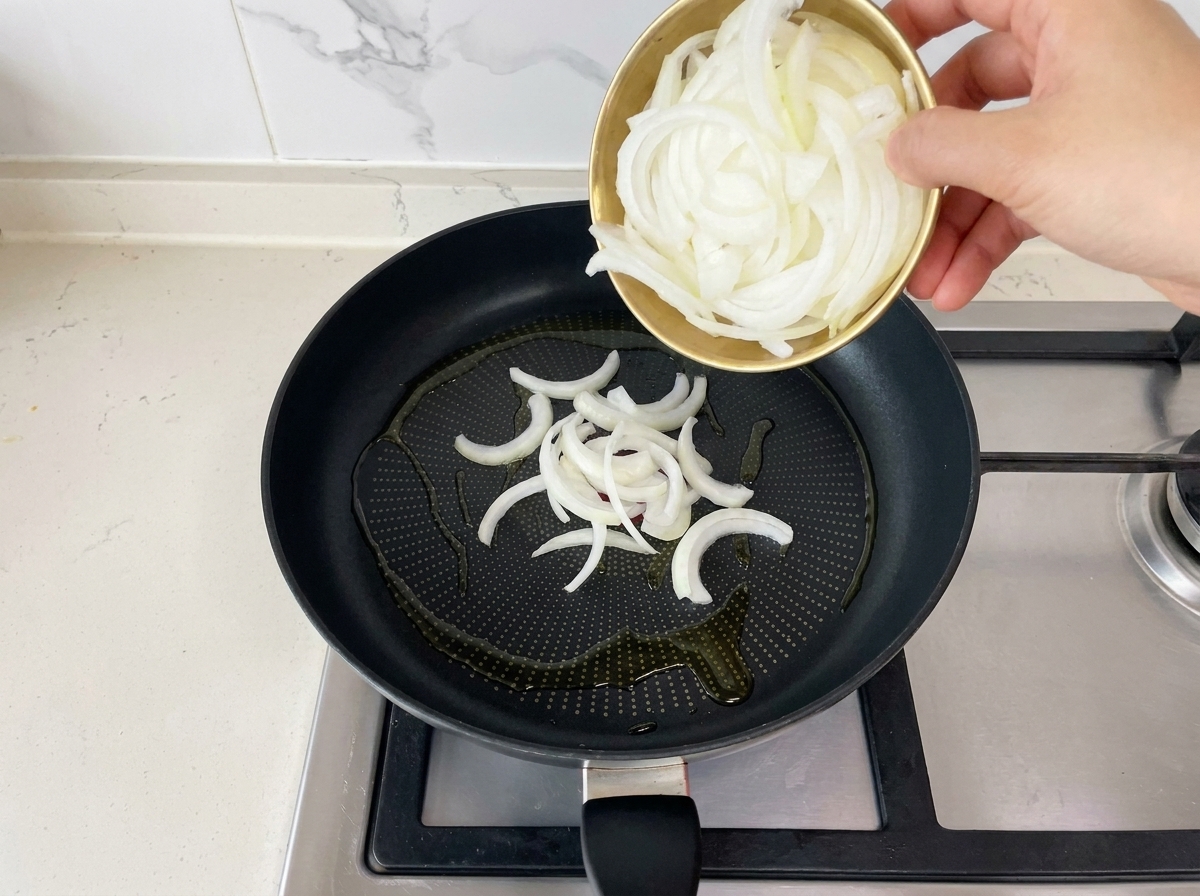 Pouring freshly sliced white onions from a small gold bowl into a black frying pan containing hot oil.