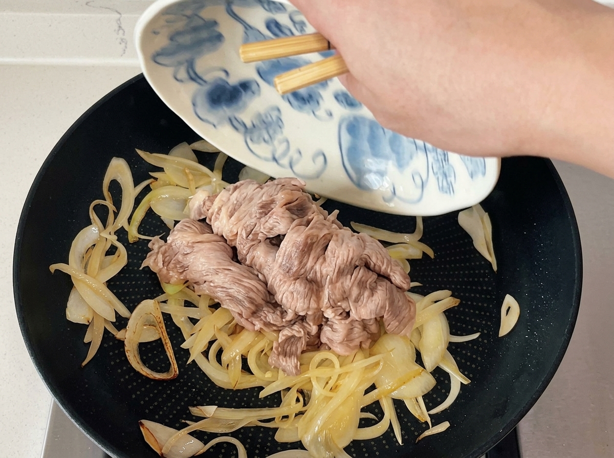 Chopsticks sliding blanched beef slices from a patterned plate into a pan of sautéed onions.