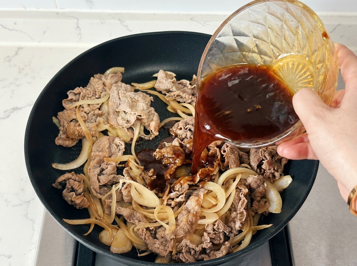 Hand pouring a dark seasoning sauce from a glass bowl over beef and onions in a black skillet.