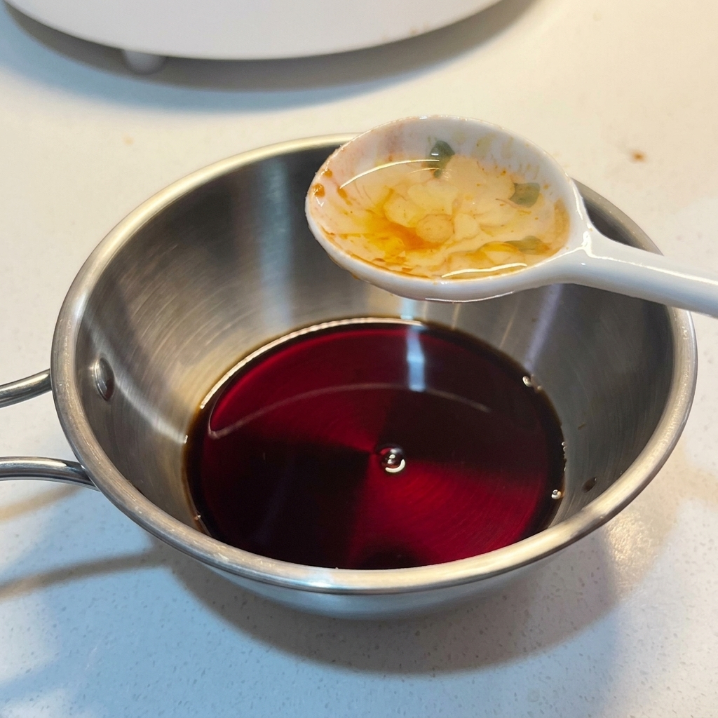 A spoonful of liquid seasoning being added into a small metal bowl containing a dark sauce mixture.