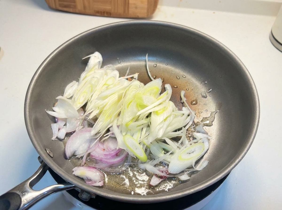 Sliced white onions and scallions sizzling in hot oil in a metal skillet.