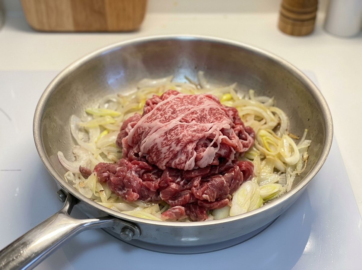 A pile of raw, thinly sliced red fatty beef resting on top of cooked onions in a metal skillet.