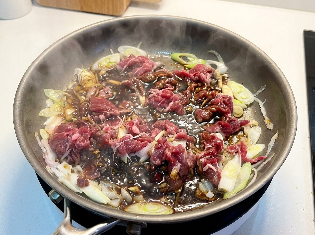 Partially cooked sliced beef and onions sitting in a dark, rich simmering sauce in a skillet.