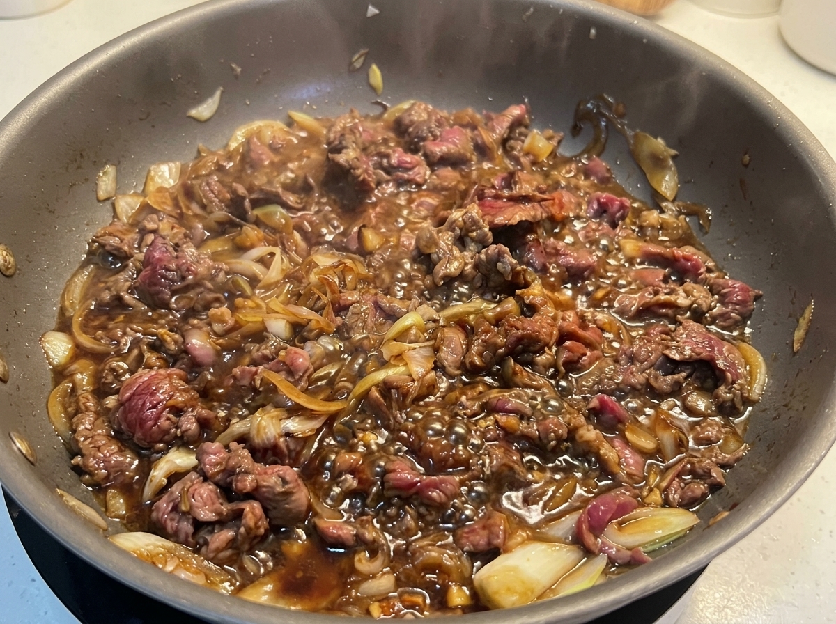Fully browned thinly sliced beef simmering vigorously in a bubbling dark sauce with onions.