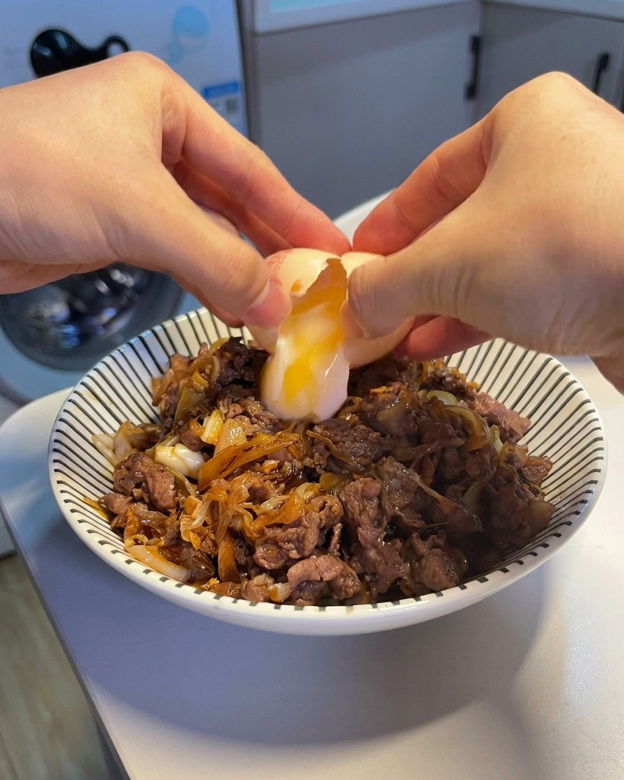 Two hands cracking an onsen egg, dropping a soft, runny yolk and white onto a steaming beef and rice bowl.