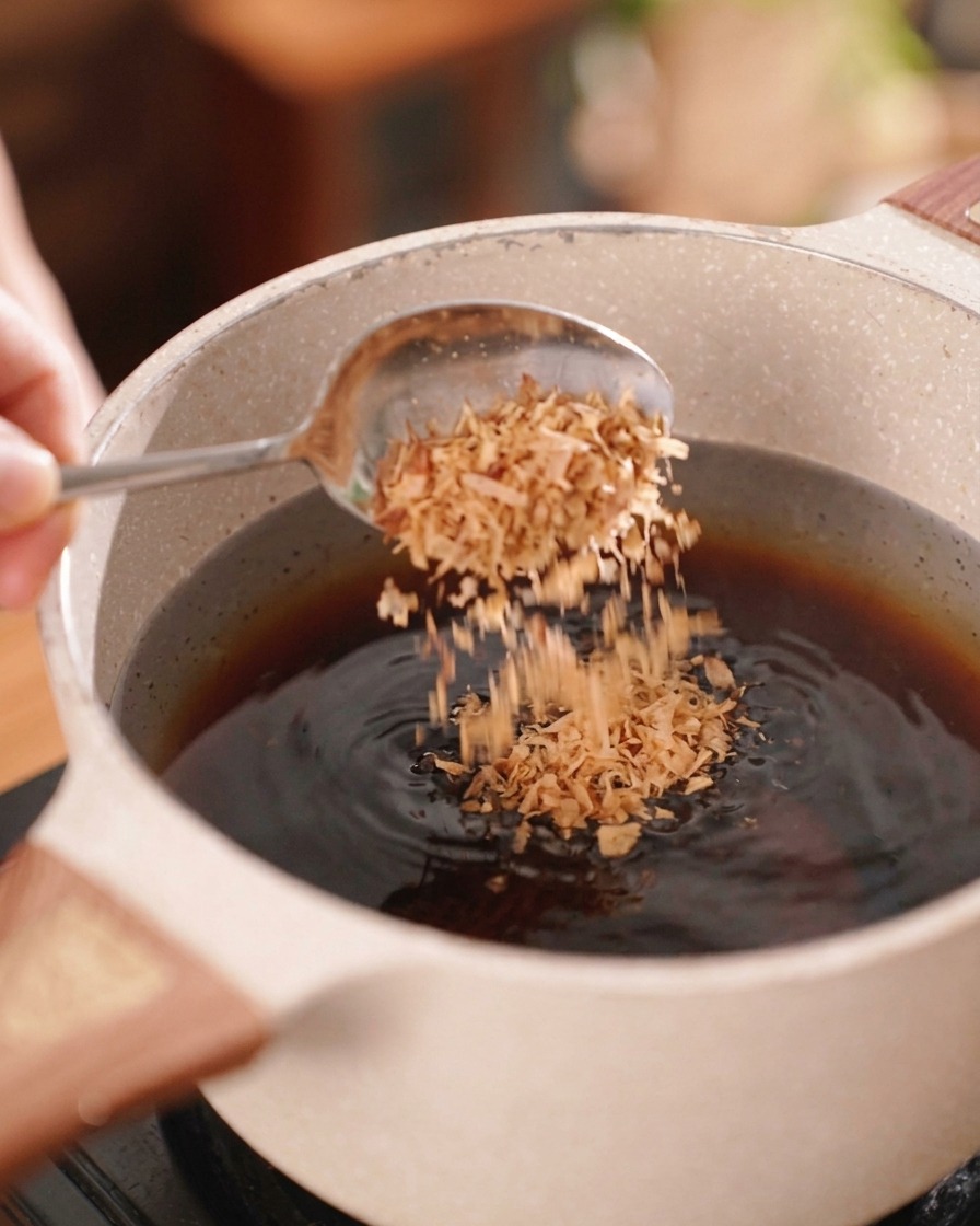 A person using a silver spoon to sprinkle crispy brown fried shallots into a pot of dark savory marinade.