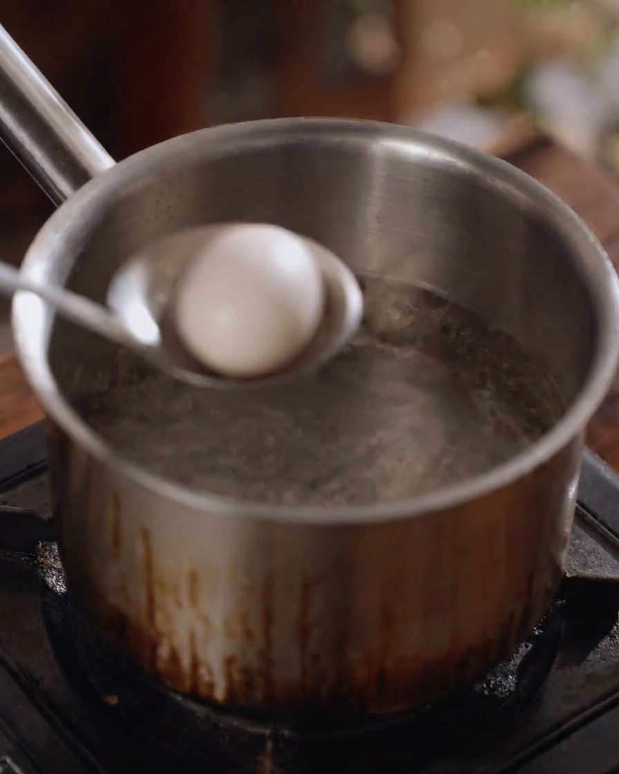 A whole white egg being gently lowered into a pot of boiling water with a metal ladle.
