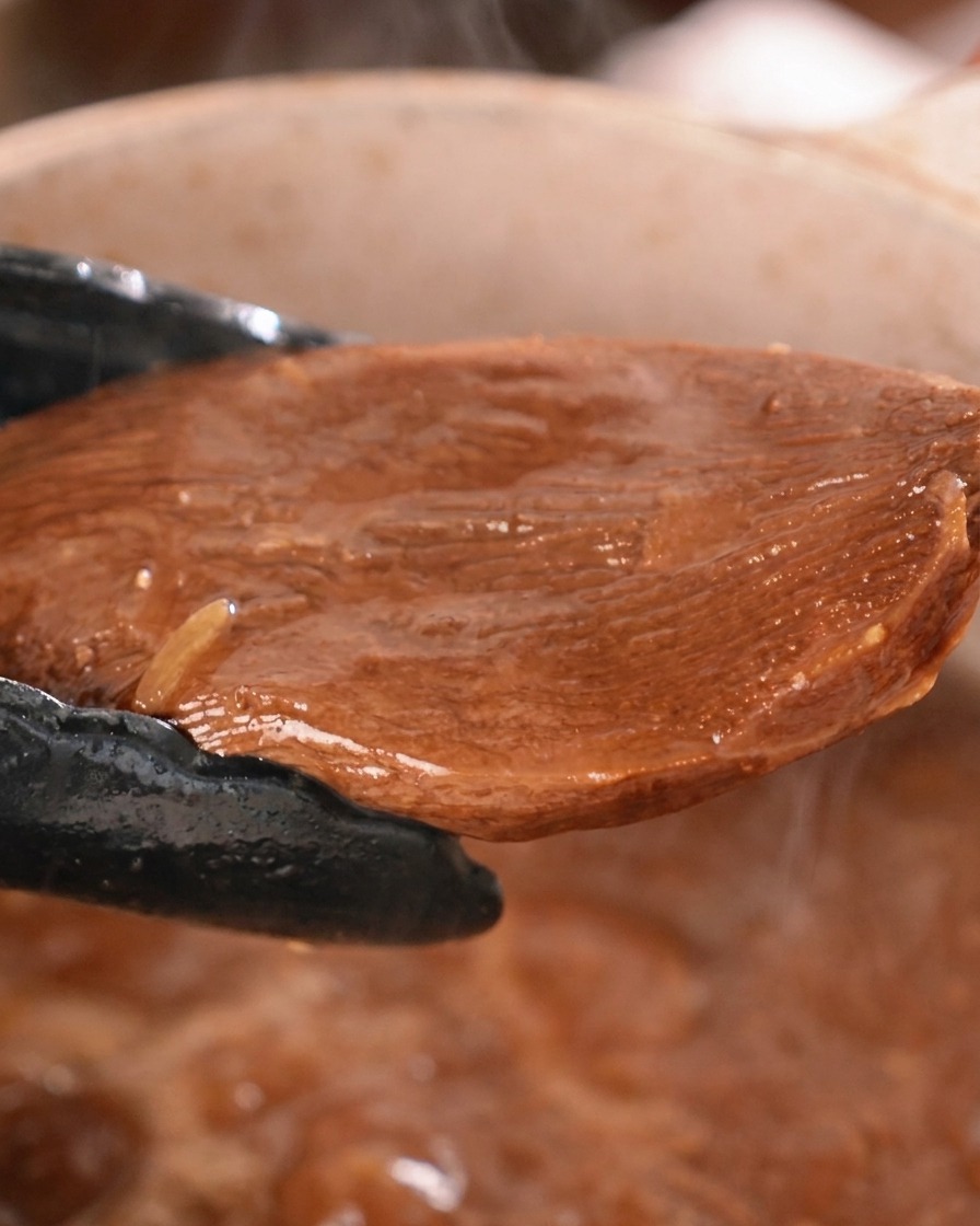 A thick piece of braised beef tongue being lifted from a pot of dark sauce with black kitchen tongs.
