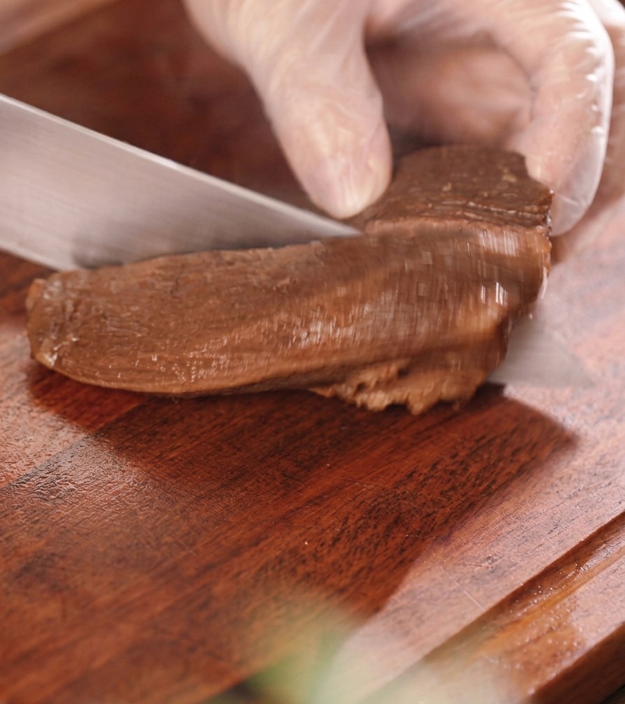 A gloved hand slicing a piece of dark, braised beef tongue into thick bite-sized pieces on a wooden board.