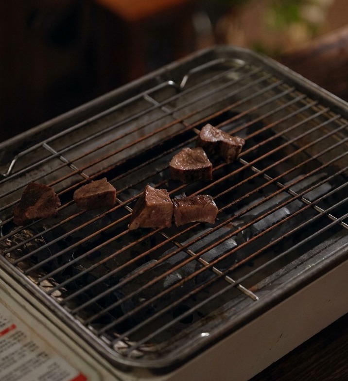 Slices of braised beef tongue resting on a small portable wire grill grate over charcoal.