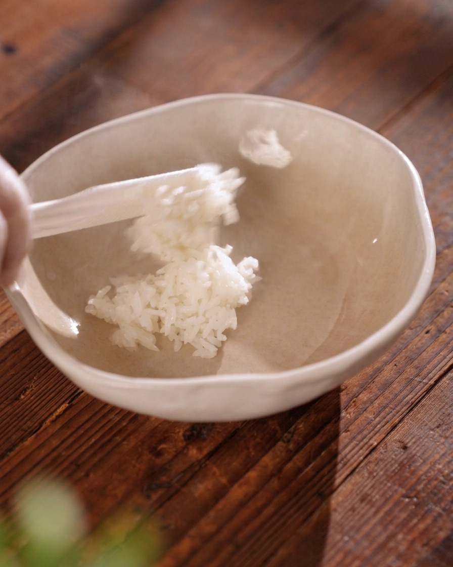 A white rice paddle transferring fluffy, steaming white rice into a light beige ceramic bowl.