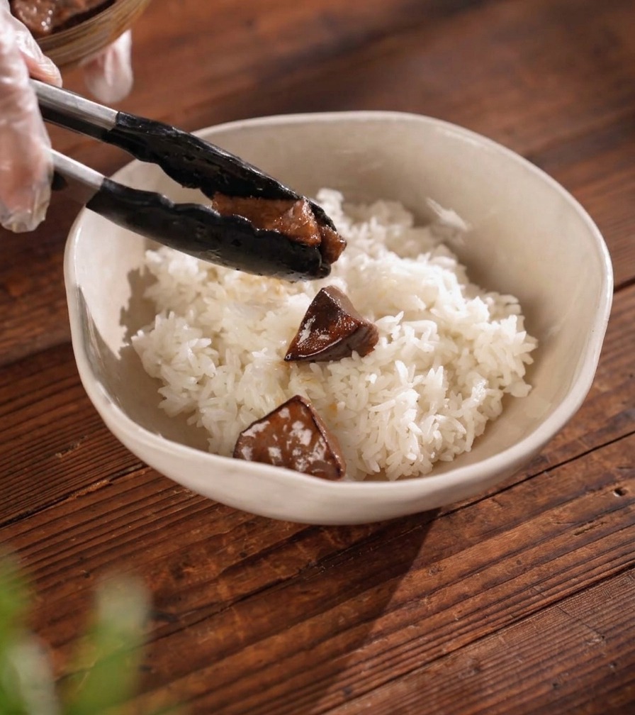 Tongs placing a piece of dark-glazed, grilled meat onto a bed of fluffy white rice in a ceramic bowl.