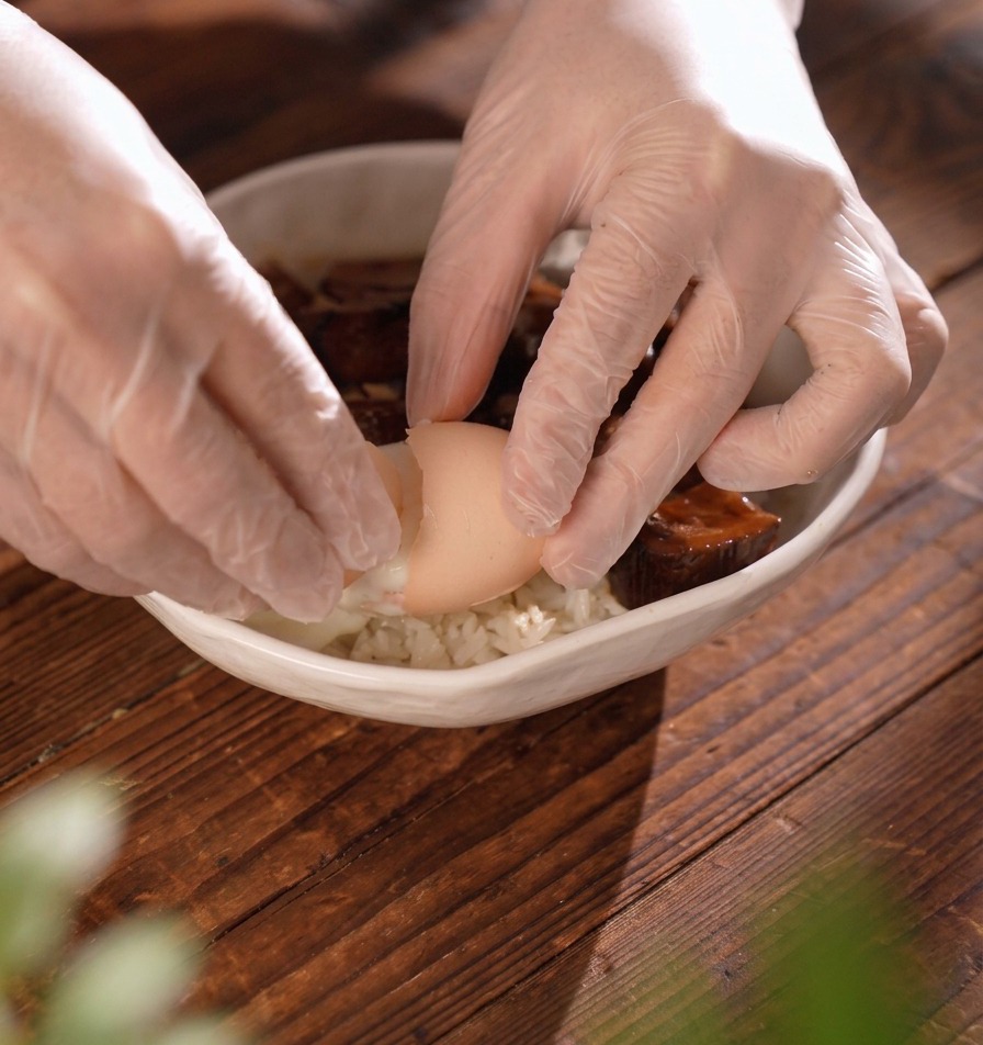 Gloved hands carefully cracking a soft eggshell to release a poached egg into a rice bowl with meat.