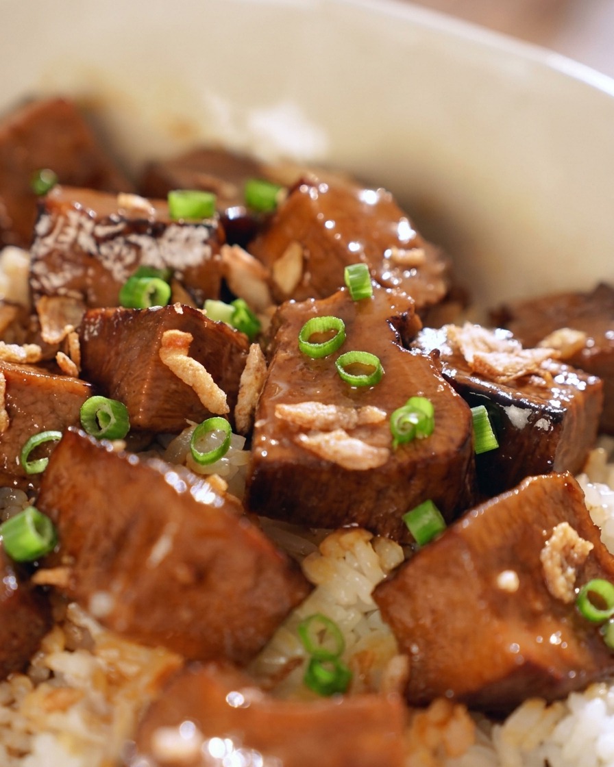 A close-up of glazed beef chunks on rice, topped with bright green scallion rings and golden-brown fried shallots.