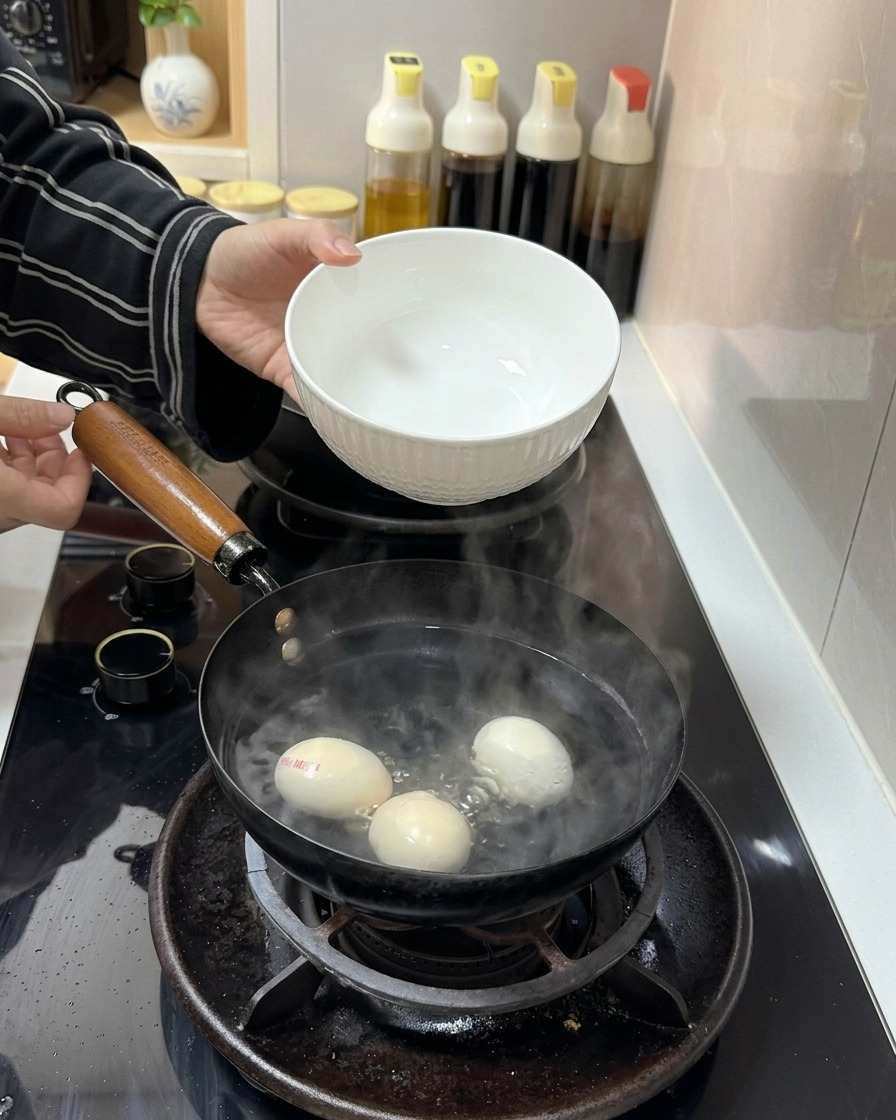 A white ceramic bowl pouring clear cold water into a pot containing boiling water and three eggs.