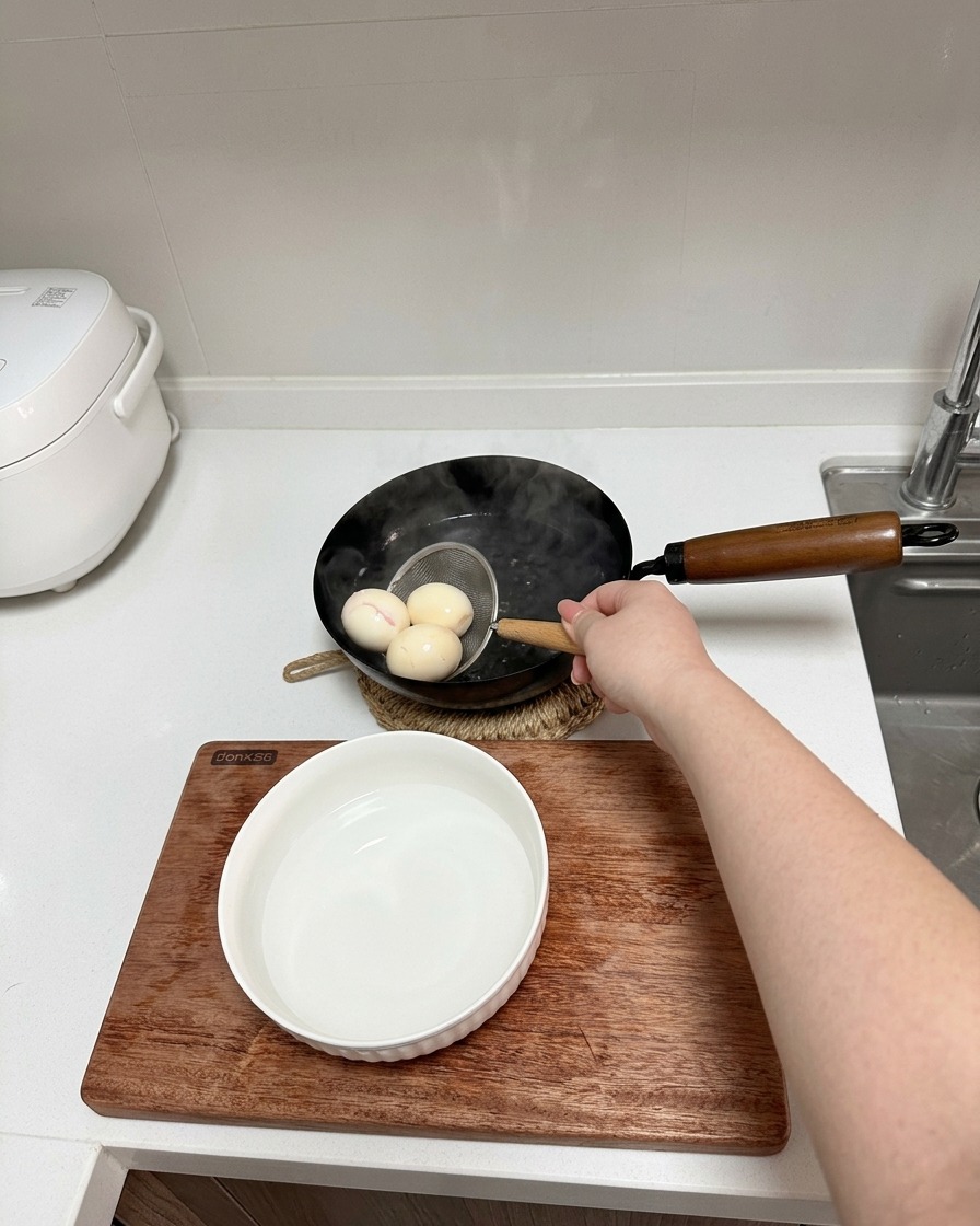 A hand using a small wire strainer to lift three boiled eggs from a pot toward a bowl of cold water.