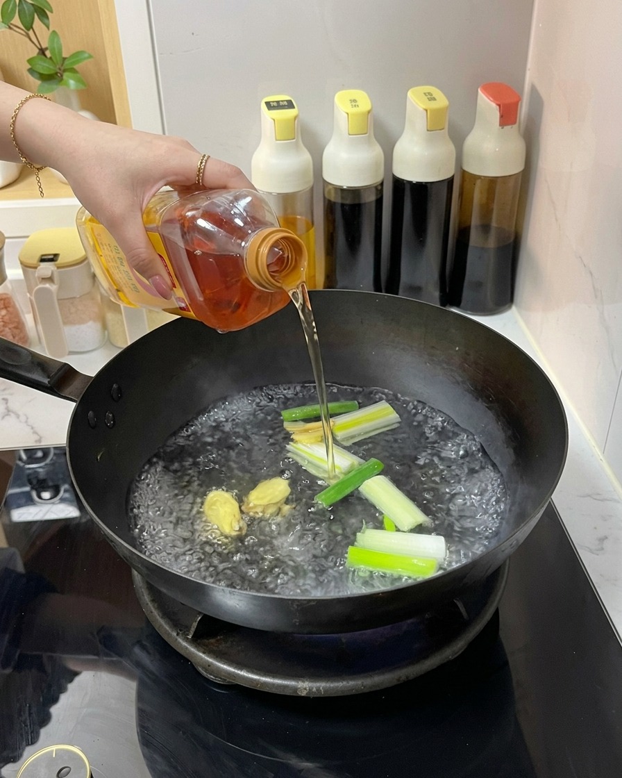 A person pouring a clear amber liquid from a plastic bottle into a wok containing boiling water, ginger, and green onions.