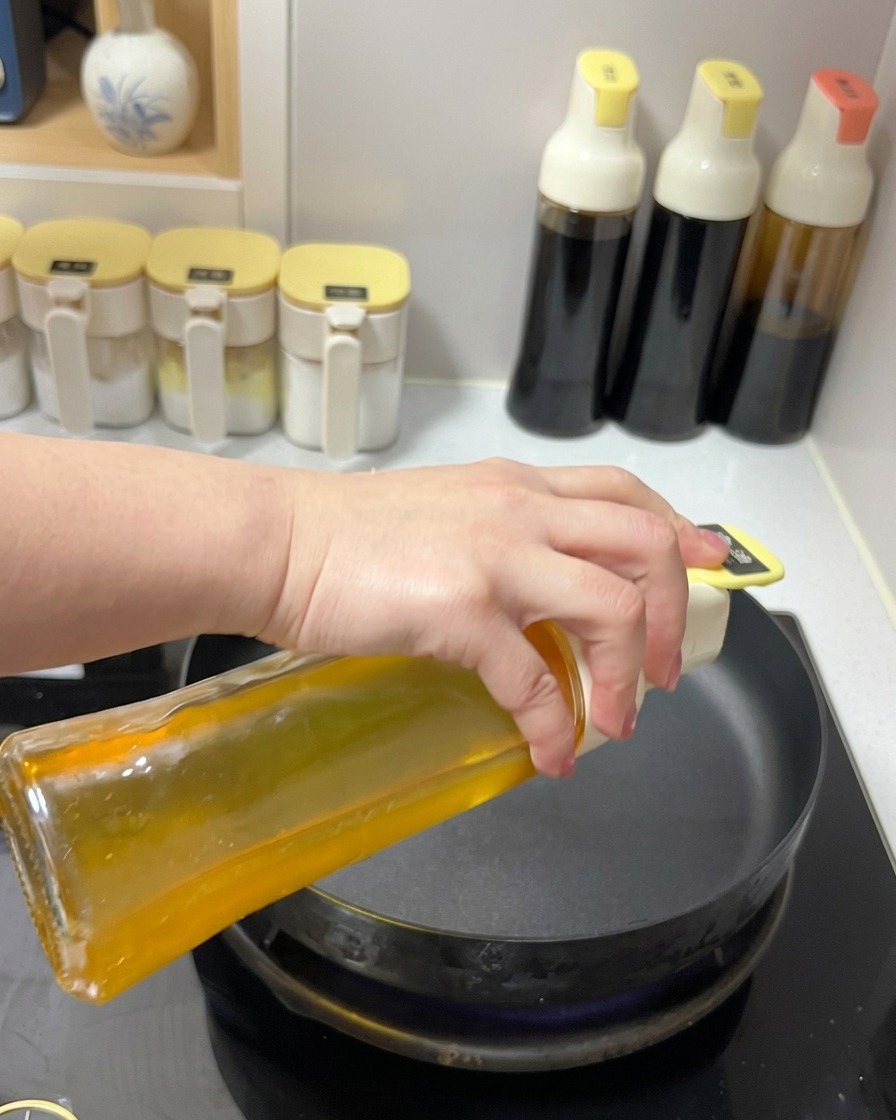 Clear cooking oil being poured from a glass dispenser into a black frying pan on a stovetop.