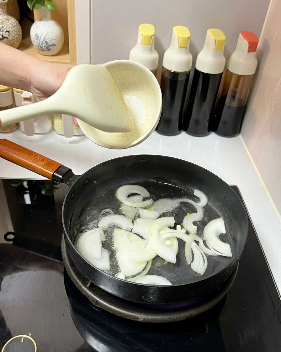 Slices of white onion being tossed in a frying pan with oil using a white spatula.