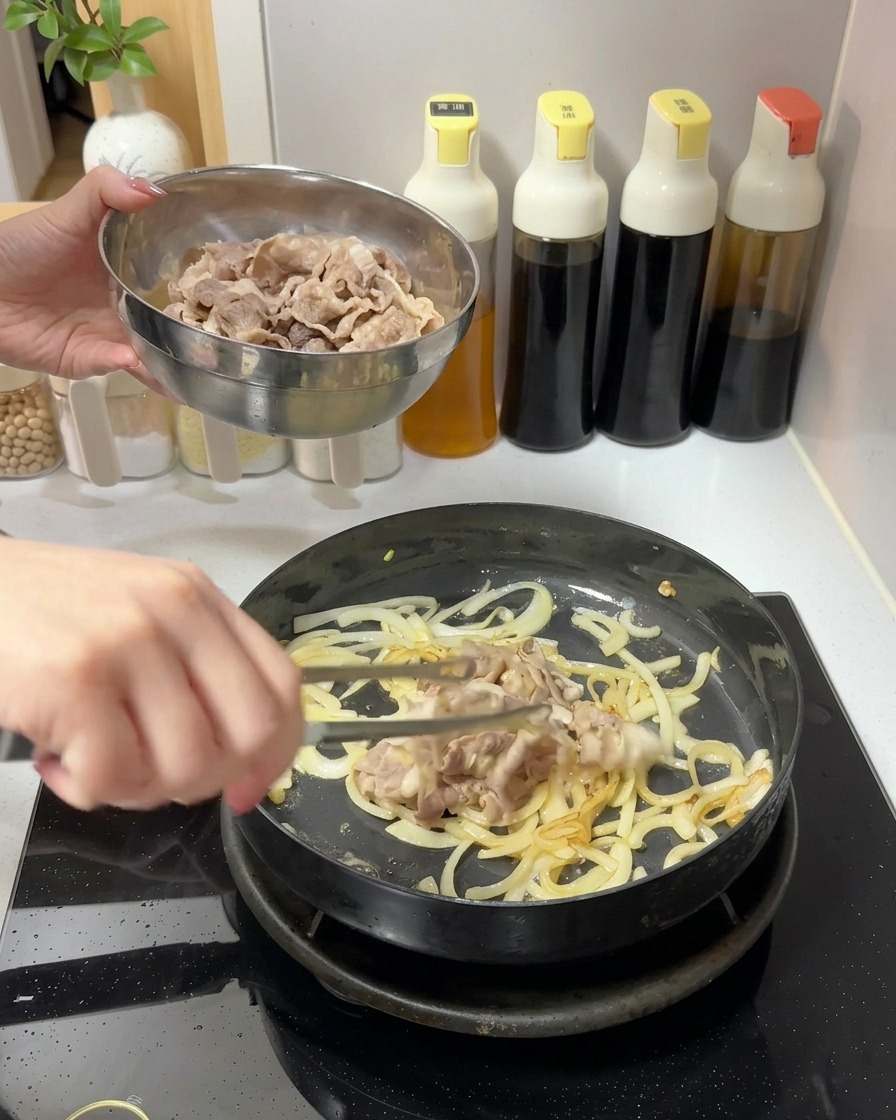 Adding a bowl of cooked beef slices into a pan containing sautéed onions.