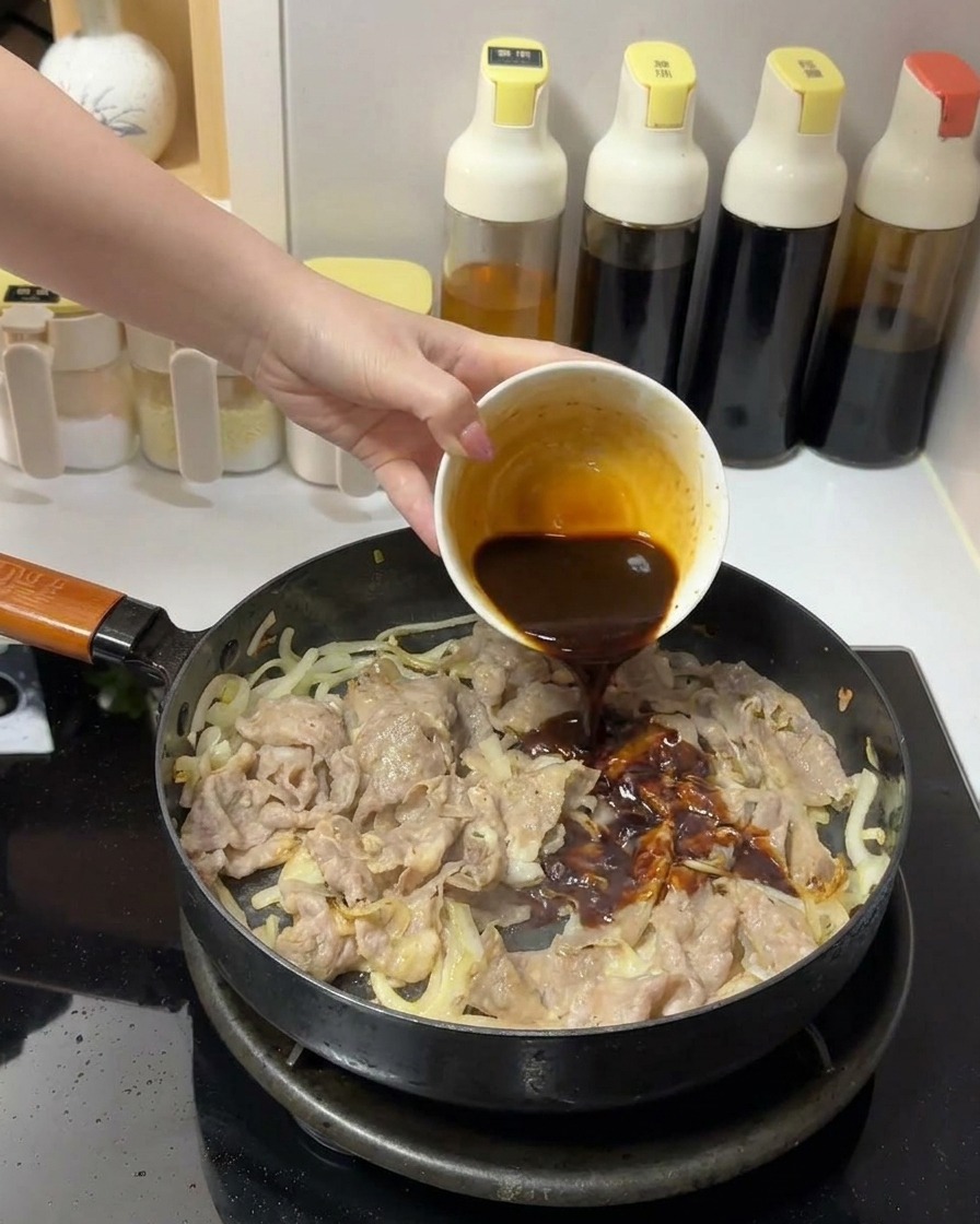 A thick, dark brown sauce being poured from a small white bowl onto beef and onions in a frying pan.
