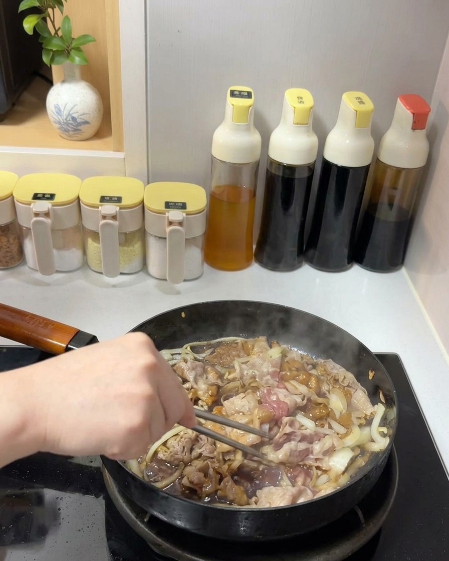 Beef and onions being tossed with a dark, glossy sauce in a pan using metal chopsticks.