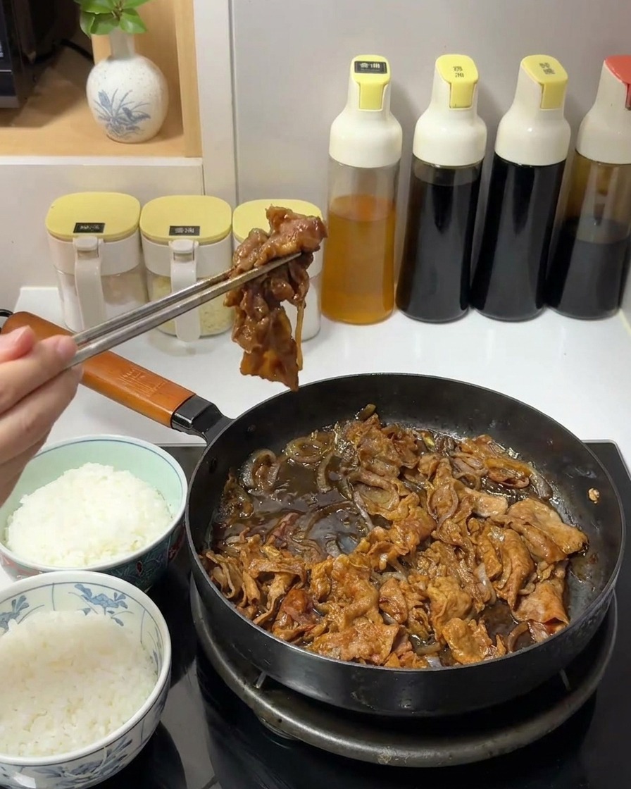 A hand using metal chopsticks to lift a portion of dark, saucy beef and onions from a pan.