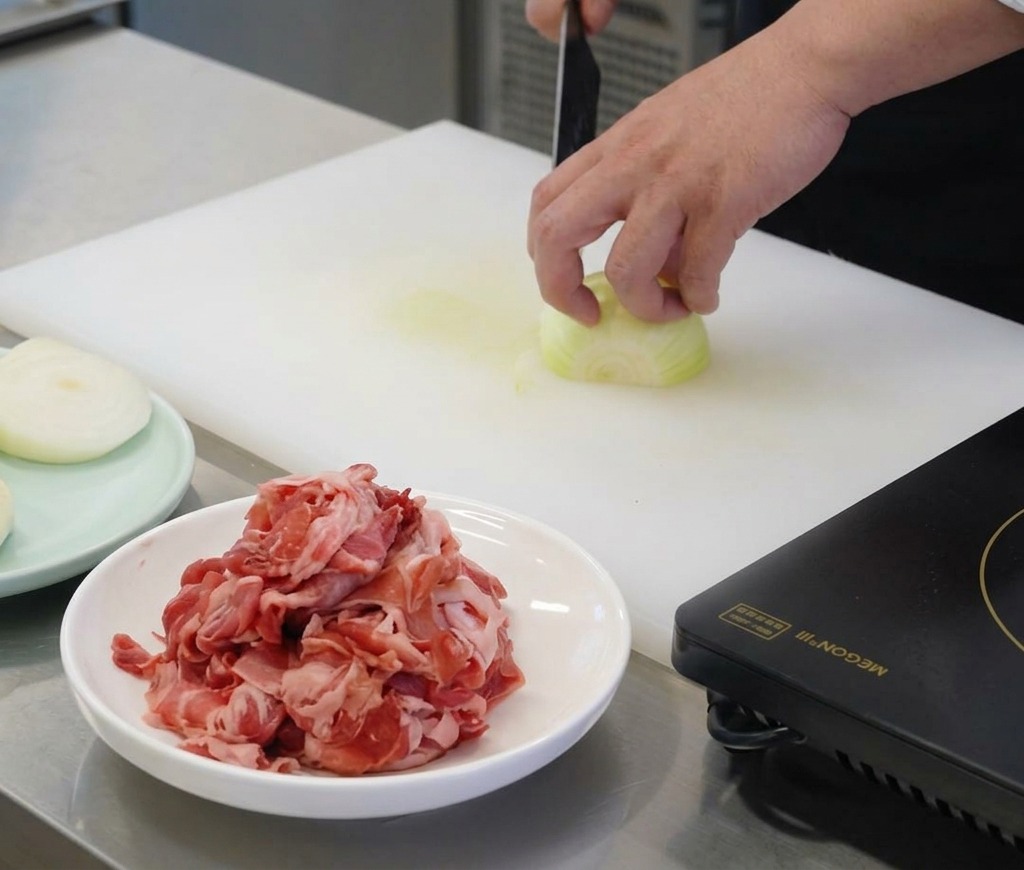 A chefs hand holding a white onion on a white cutting board while slicing it into thin vertical strips with a sharp knife.