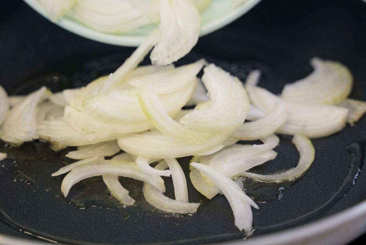Slices of raw white onion being poured from a light green plate into a black frying pan with shimmering cooking oil