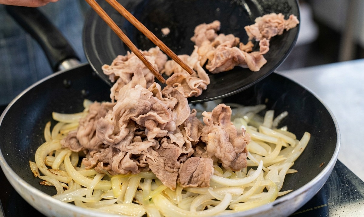 A person using wooden chopsticks to slide blanched beef strips from a black plate into a pan containing sautéed onions