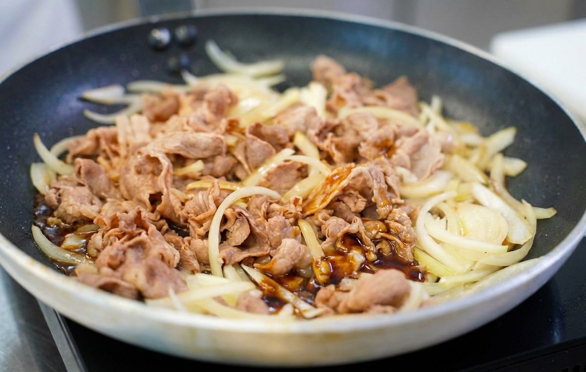 A dark, glossy brown savory sauce being poured over a mixture of stir-fried beef and onions in a frying pan.