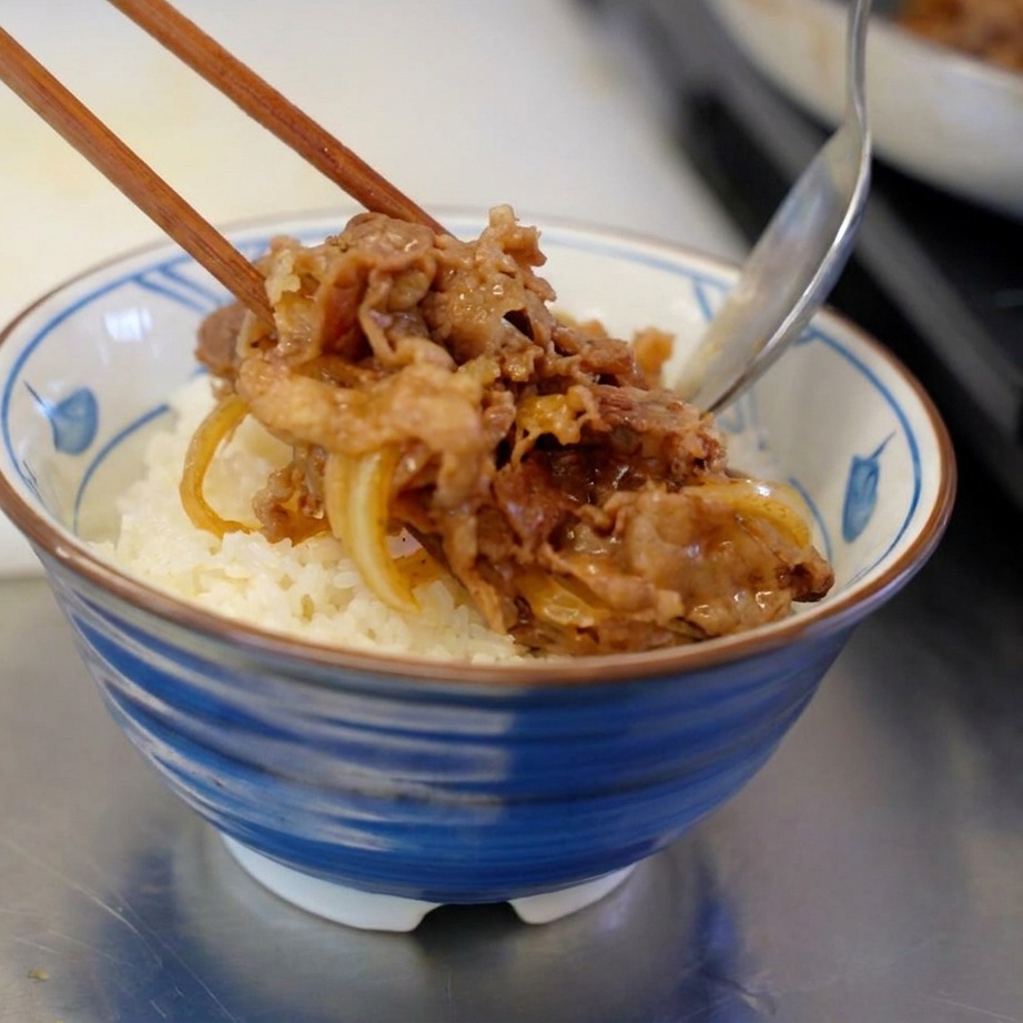 A portion of savory cooked beef and onions being placed onto a bowl of fluffy white rice using chopsticks and a spoon.