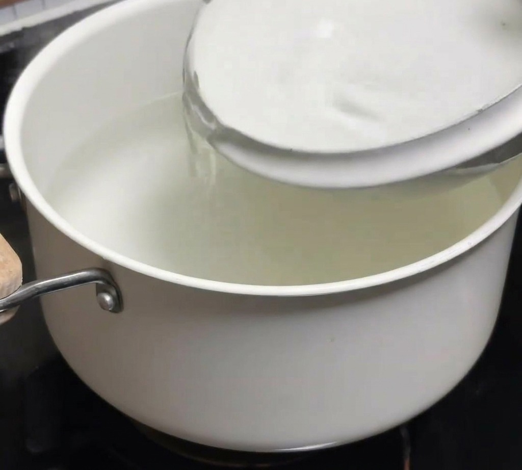 A bowl of cold water being poured into a pot of recently boiled hot water.