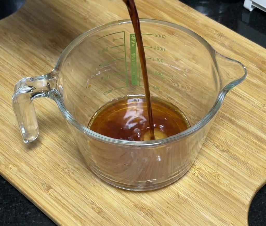 Dark soy sauce being poured into a glass measuring cup containing honey and seasonings.