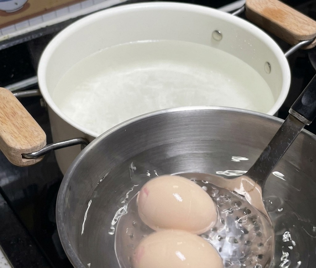 Two eggs resting in a stainless steel bowl filled with ice water, with a white pot used for soaking visible in the background.