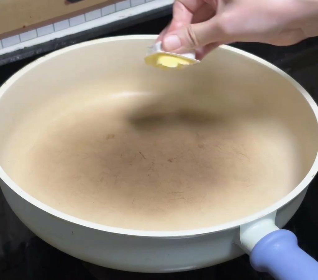 A hand placing a square of yellow butter into a clean white ceramic frying pan on a stovetop.