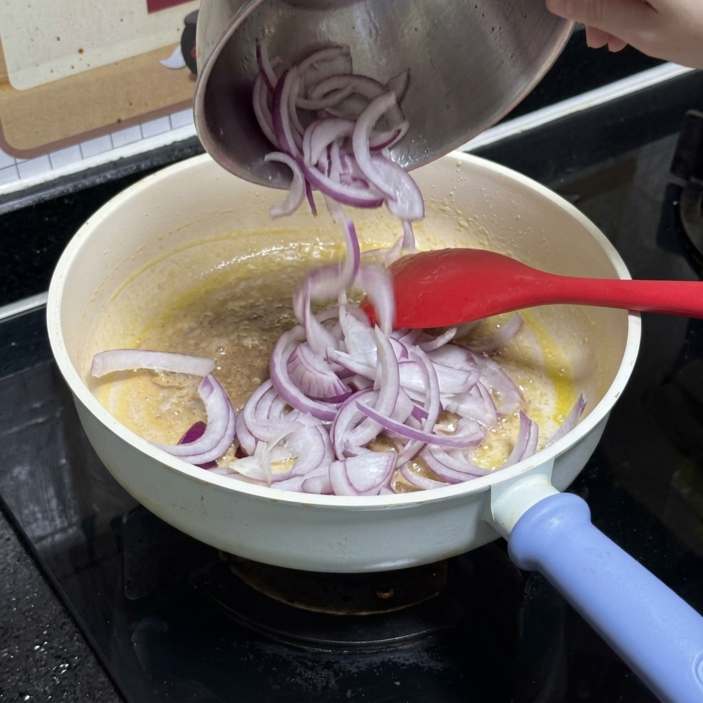 Sliced red onions being poured from a stainless steel bowl into a white frying pan containing melted butter.